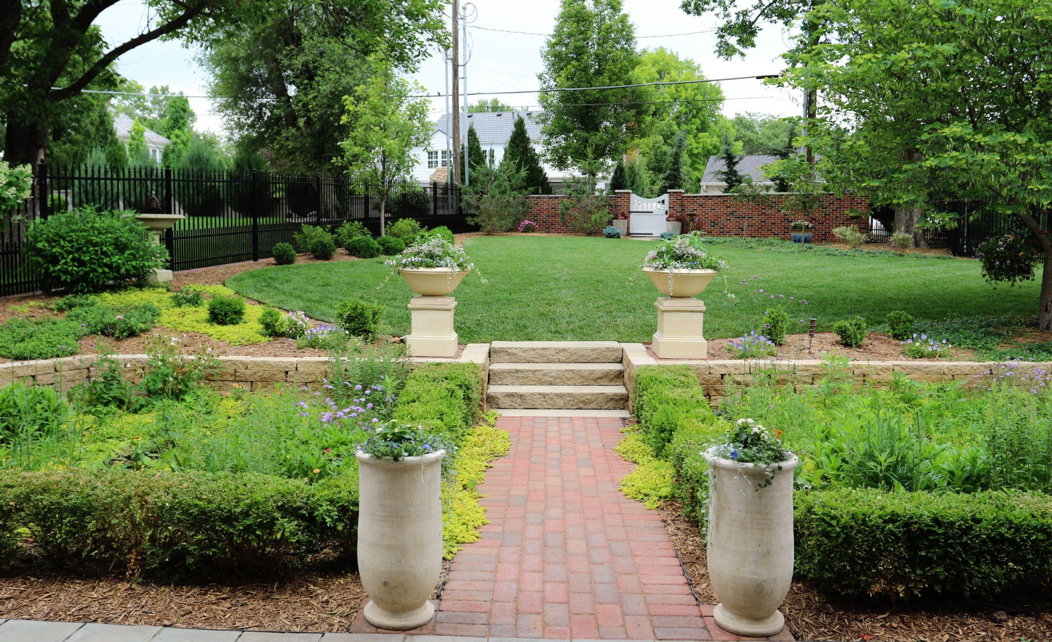 A brick pathway leading through a formal garden with boxwood hedges, flowering plants, stone planters, and steps up to a grassy lawn enclosed by fencing.