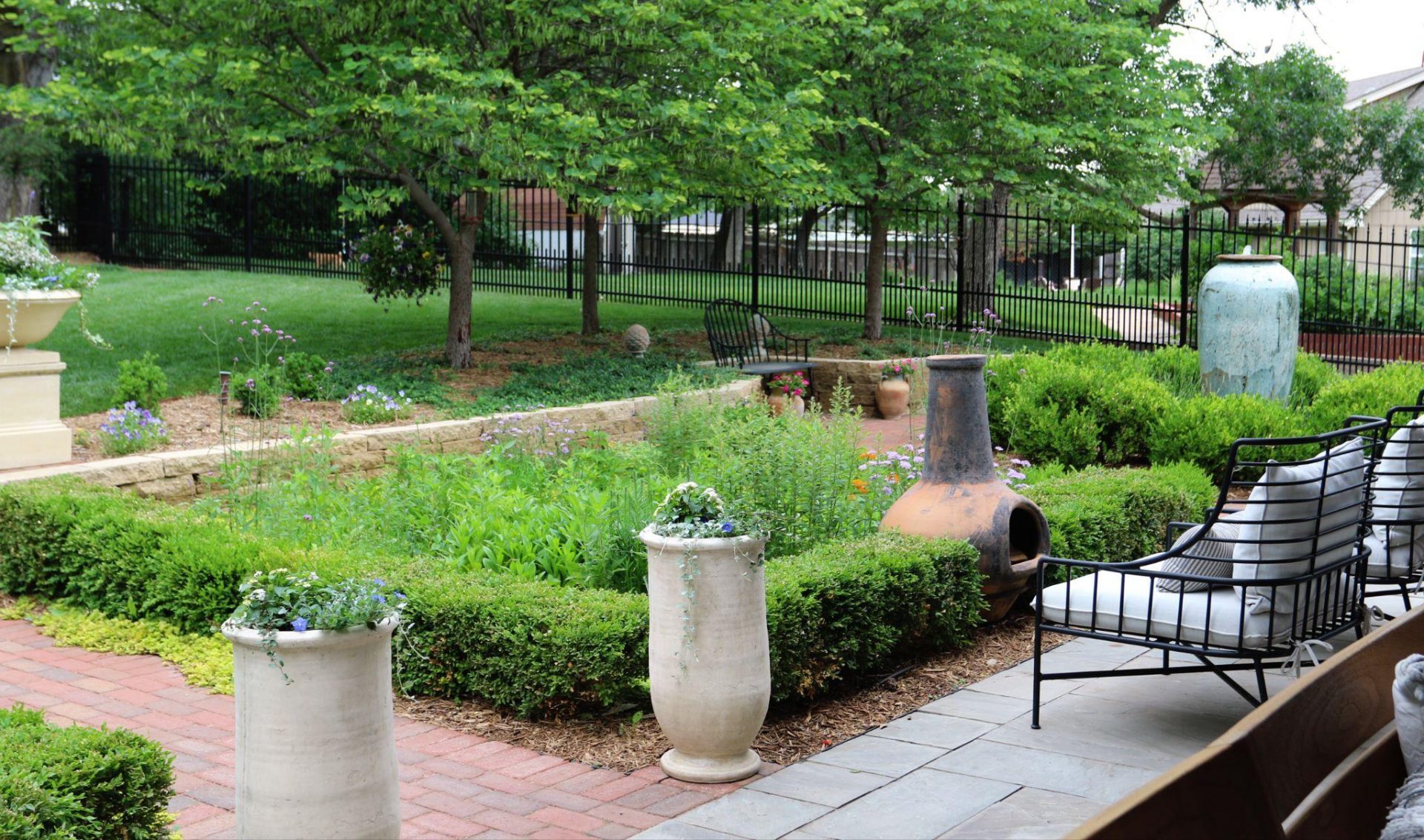 A garden seating area with cushioned metal chairs, tall ceramic planters, boxwood hedges, flowering plants, and mature trees creating a lush outdoor space.
