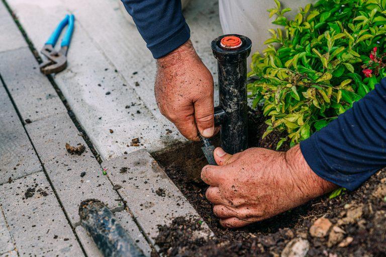Close-up of a worker’s hands repairing an irrigation sprinkler head next to a garden bed.