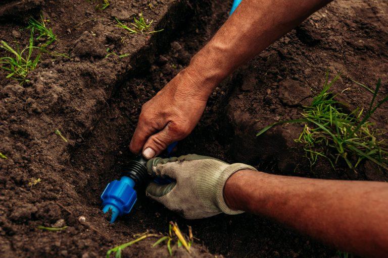 Hands connecting a blue irrigation pipe fitting inside a freshly dug trench.
